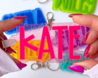 以下が含まれることがあります： 「JOEL」、「WILEY」、「KATE」、「SADIE」、そしてもう1つの名前（不明瞭）が書かれた、カラフルでパーソナライズされたキーホルダーのセット。キーホルダーは、キラキラ光る透明なプラスチック製で、3D効果があります。