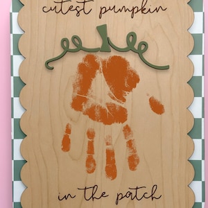 May include: Wooden sign with a scalloped edge and a green and white checkered border. The sign features a handprint in orange, with the words "cutest pumpkin in the patch" and the name "ADRIAN 2025" in a script font.