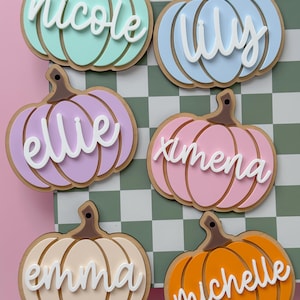 May include: A collection of colorful pumpkin-shaped name tags. Each tag features a different name in white script: Nicole, Lily, Ellie, Ximena, Emma, and Michelle. The pumpkins are in shades of teal, blue, purple, pink, cream, and orange.