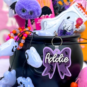 Puede incluir: Un caldero negro lleno de art&iacute;culos de tem&aacute;tica de Halloween, que incluyen un murci&eacute;lago peludo morado, un fantasma blanco y un collar de cuentas rosa y naranja. Un lazo de purpurina morado con el nombre "Addie" est&aacute; unido al caldero.