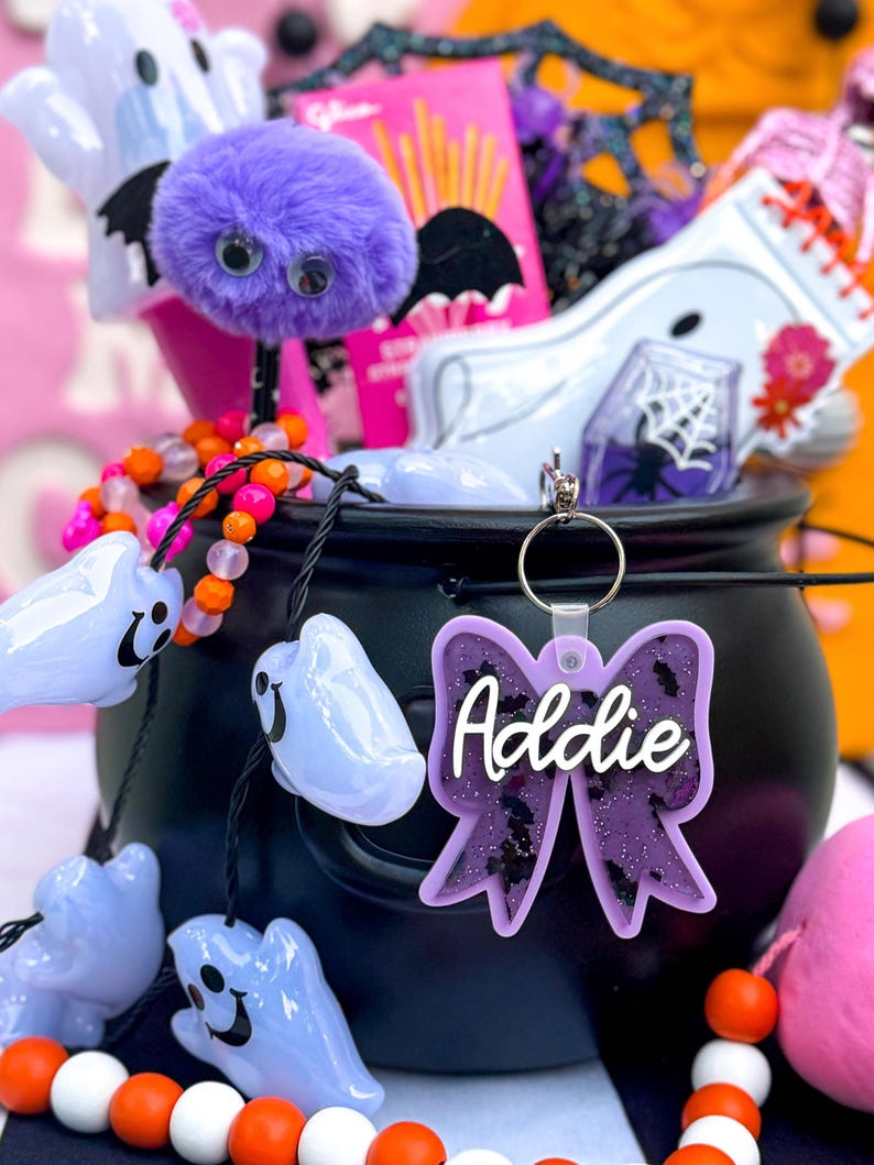 Puede incluir: Un caldero negro lleno de art&iacute;culos de tem&aacute;tica de Halloween, que incluyen un murci&eacute;lago peludo morado, un fantasma blanco y un collar de cuentas rosa y naranja. Un lazo de purpurina morado con el nombre "Addie" est&aacute; unido al caldero.