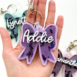 Puede incluir: Llavero de lazo de resina con purpurina morada con el nombre "Addie" en letras blancas. El llavero tiene un anillo de metal plateado y un clip de metal plateado.