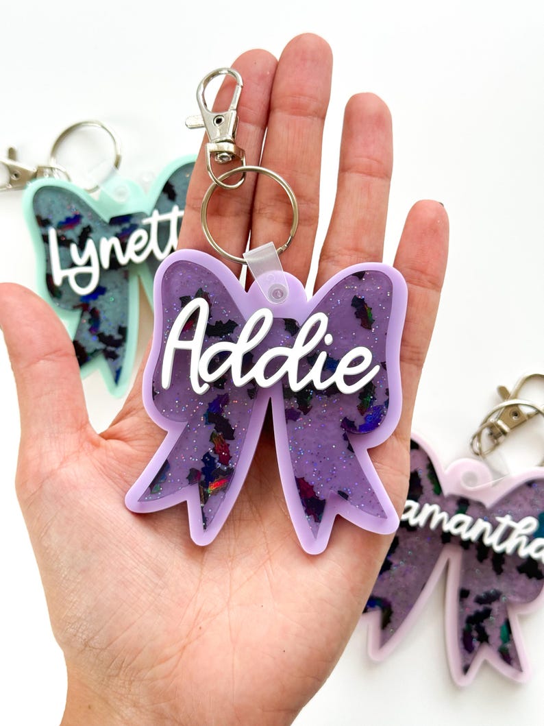Puede incluir: Llavero de lazo de resina con purpurina morada con el nombre "Addie" en letras blancas. El llavero tiene un anillo de metal plateado y un clip de metal plateado.