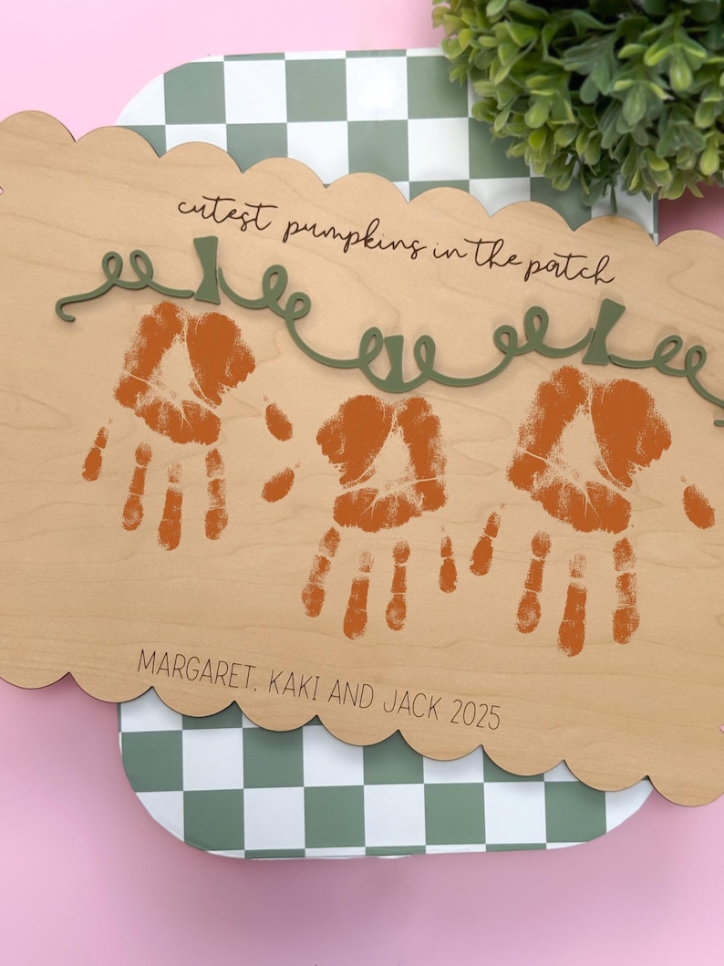 May include: Wooden sign with scalloped edges, featuring three orange handprints resembling pumpkins. The sign includes the text "cutest pumpkins in the patch" and the names "Margaret, Kaki and Jack 2025" in a decorative font. A green vine design accents the top.