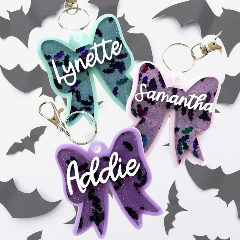 Puede incluir: Tres llaveros de lazo de resina de color morado, rosa y azul claro con purpurina y un patr&oacute;n de murci&eacute;lago negro. Cada llavero tiene un anillo de metal plateado y un cierre de metal plateado. Los llaveros est&aacute;n personalizados con los nombres "Addie", "Samantha" y "Lynette".