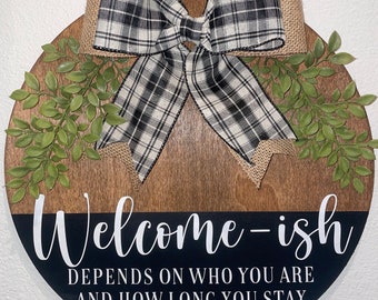 May include: Round wooden sign with the text "Welcome-ish" and the phrase "Depends on who you are and how long you stay." The sign has a black and white plaid bow, burlap, and green leaves.