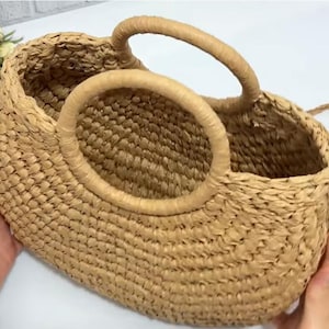 Może przedstawiać: Tkana torba typu tote ze słomy w jasnobrązowym kolorze, z dwoma uchwytami i okrągłym otworem. Torba wydaje się być wykonana z naturalnych włókien. Uchwyty są zaokrąglone i solidne, odpowiednie do przenoszenia przedmiotów.