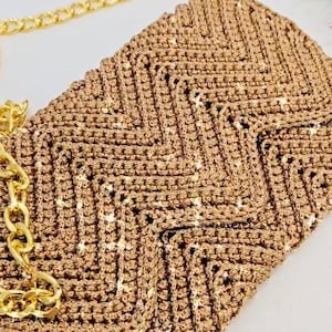 May include: A handmade, brown crochet clutch with a gold chain strap. The clutch features a textured, geometric pattern and a gold chain strap. The bag is a neutral color, suitable for various outfits and occasions.