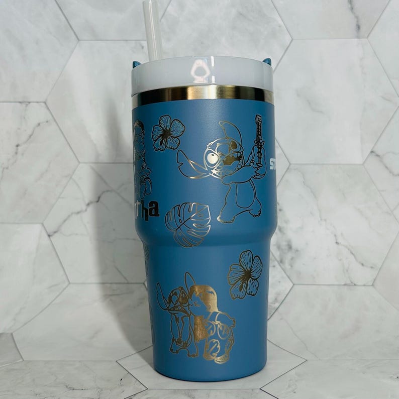 May include: A blue stainless steel tumbler with a white lid and two straws. The tumbler features a gold design of Stitch from Lilo & Stitch, along with tropical flowers and leaves. The text "Aloha" is also included in the design.