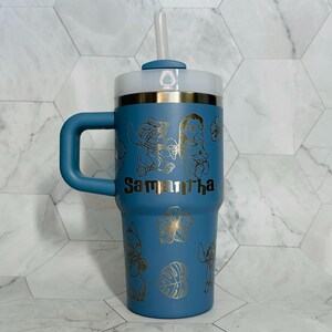 May include: A blue stainless steel travel mug with a handle and a straw. The mug has a gold design of Stitch and Angel from Lilo & Stitch, along with tropical flowers and leaves. The name "Samantha" is engraved on the mug.