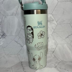 Peut inclure: Un gobelet isotherme en acier inoxydable vert clair avec un motif graphique noir et blanc représentant Stitch et Lilo du film Lilo & Stitch de Disney. Le gobelet est doté d'un couvercle noir avec une poignée de transport et le logo Stanley est imprimé sur le côté.