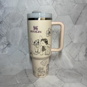 May include: A pink stainless steel travel mug with a black and white graphic of Stitch and Angel from the Disney movie Lilo & Stitch. The mug has a black handle and a black Stanley logo.