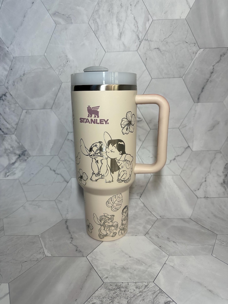 May include: A pink stainless steel travel mug with a black and white graphic of Stitch and Angel from the Disney movie Lilo & Stitch. The mug has a black handle and a black Stanley logo.