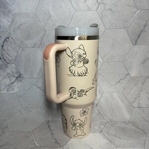 May include: A light pink stainless steel travel mug with a black handle and a white lid. The mug features a black and white graphic of Stitch from Lilo & Stitch. The graphic includes Stitch playing the ukulele and surrounded by flowers.