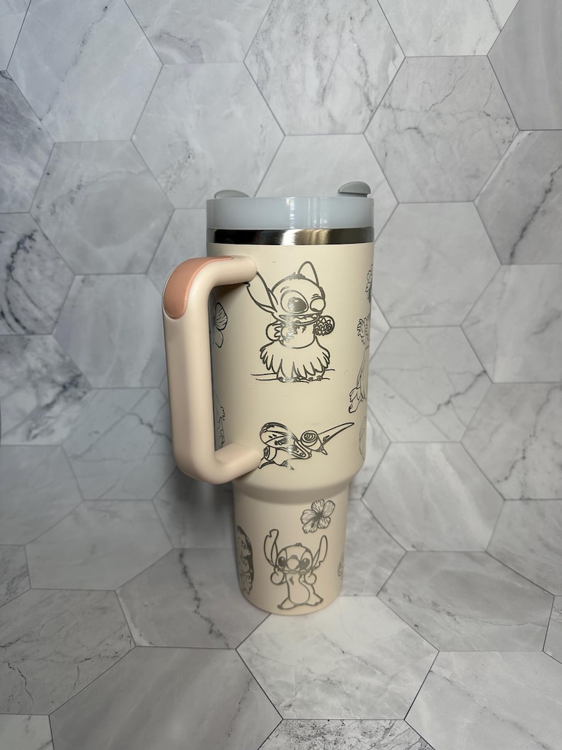 May include: A light pink stainless steel travel mug with a black handle and a white lid. The mug features a black and white graphic of Stitch from Lilo & Stitch. The graphic includes Stitch playing the ukulele and surrounded by flowers.