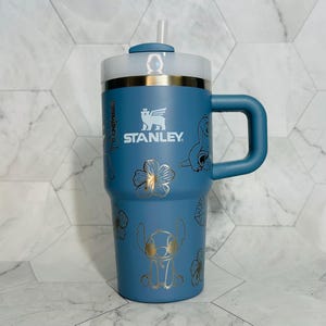 May include: Blue stainless steel travel mug with a lid and handle. The mug features a gold design of Stitch from Lilo & Stitch, flowers, and the Stanley logo.