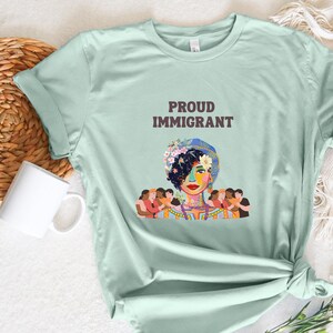 May include: A light green t-shirt with the text "PROUD IMMIGRANT" printed on the front. The shirt features a colorful illustration of a woman with flowers in her hair and people surrounding her.