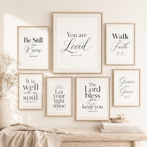 May include: A set of framed art prints featuring motivational quotes in black script on a white backdrop. The prints include phrases such as "Be Still and Know," "You are Loved," and "Walk by Faith." The frames are light wood, and the prints are displayed on a wooden surface.
