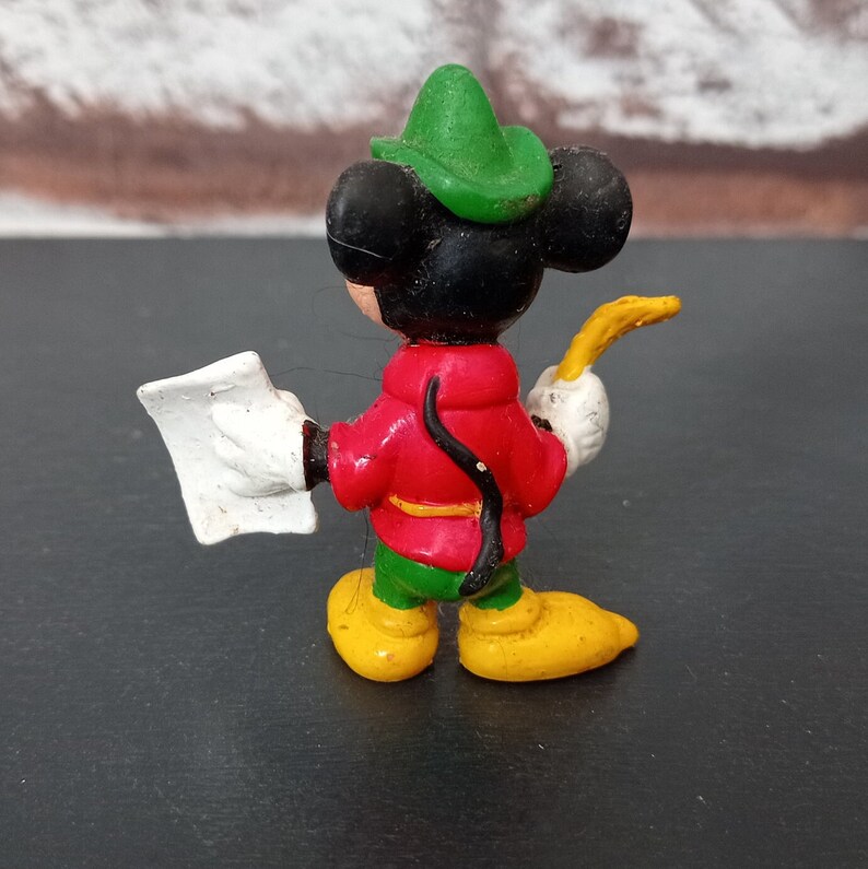 May include: A plastic figurine of Mickey Mouse dressed as Robin Hood. He is wearing a green hat, red shirt, green pants, and yellow shoes. He is holding a yellow horn and a white piece of paper.
