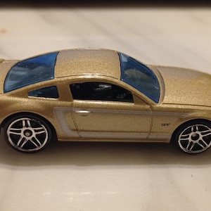 May include: A gold toy car model of a Ford Mustang GT. The car has black wheels and a blue tinted windscreen.