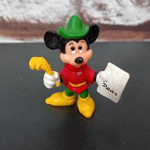May include: A plastic figurine of Mickey Mouse dressed as a forest ranger. He is wearing a green hat, red shirt, green pants, and yellow boots. He is holding a yellow stick in one hand and a white paper with the word "Dear" written on it in the other hand.