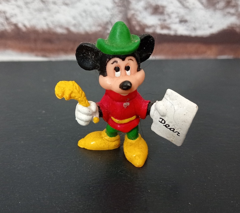 May include: A plastic figurine of Mickey Mouse dressed as a forest ranger. He is wearing a green hat, red shirt, green pants, and yellow boots. He is holding a yellow stick in one hand and a white paper with the word "Dear" written on it in the other hand.