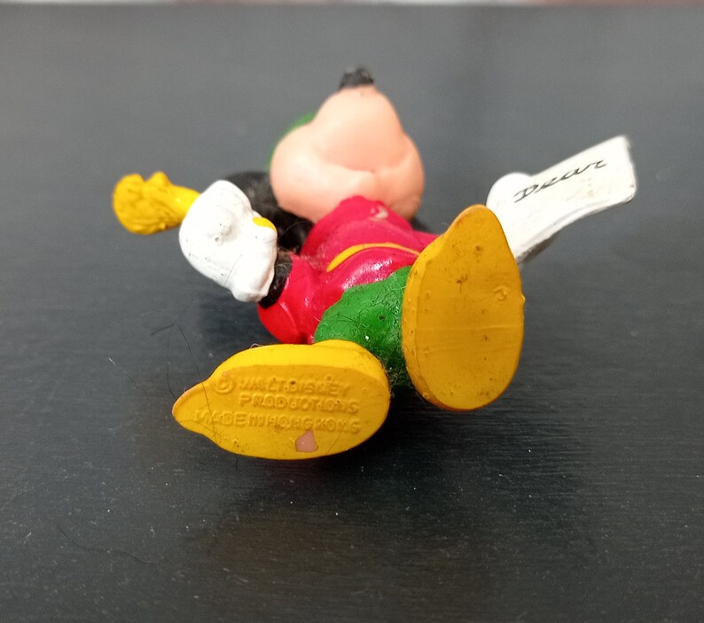 May include: A yellow, red, green, and white plastic Mickey Mouse figurine holding a paper with the word "Dear" written on it. The figurine is made by Walt Disney Productions and is made in Hong Kong.