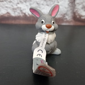 May include: A gray and white rabbit figurine with pink inner ears and a brown nose. The rabbit has a white collar and a tag with text. The figurine is on a black surface.