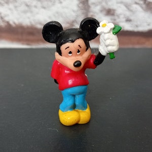 Puede incluir: Una peque&ntilde;a figura de pl&aacute;stico de Mickey Mouse sosteniendo una flor blanca. Mickey lleva una camisa roja, pantalones azules y zapatos amarillos. Tiene una nariz negra y ojos negros.