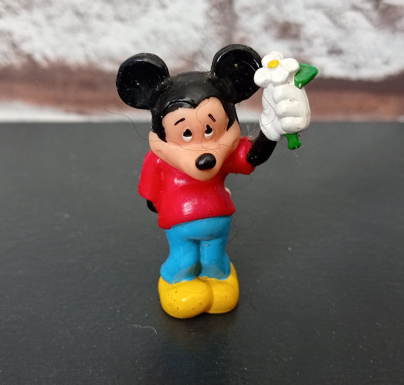 Puede incluir: Una peque&ntilde;a figura de pl&aacute;stico de Mickey Mouse sosteniendo una flor blanca. Mickey lleva una camisa roja, pantalones azules y zapatos amarillos. Tiene una nariz negra y ojos negros.