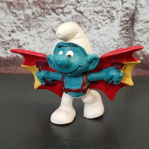 May include: A Smurf figurine with a blue body, white hat and legs, and red and yellow wings. The Smurf is smiling with outstretched arms. The figurine is on a dark surface.