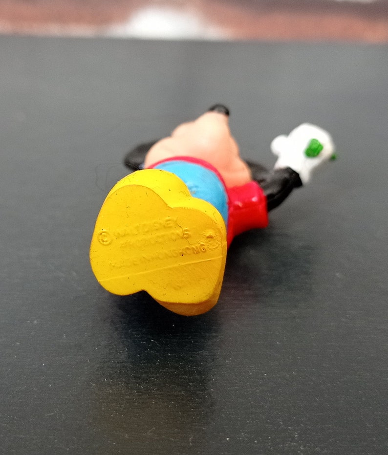 Puede incluir: Una figurita de pl&aacute;stico amarilla, roja y azul de Mickey Mouse. La figurita lleva una camisa roja, pantalones azules y zapatos amarillos. La figurita sostiene un objeto verde en su mano derecha. La parte inferior de la figurita tiene la informaci&oacute;n de copyright "&copy; Disney", "Made in China" y "Hong Kong".