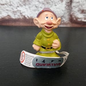 Pode incluir: Estatueta de personagem de desenho animado vestindo camisa verde, chapéu roxo e cinto preto. A estatueta tem um rosto sorridente e segura uma pulseira branca com a palavra "BULLAND" impressa.