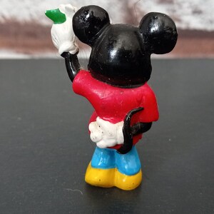 Puede incluir: Una peque&ntilde;a figura de pl&aacute;stico de Mickey Mouse con una camisa roja, pantalones azules y zapatos amarillos. Est&aacute; sosteniendo un guante blanco con una hoja verde en su mano.