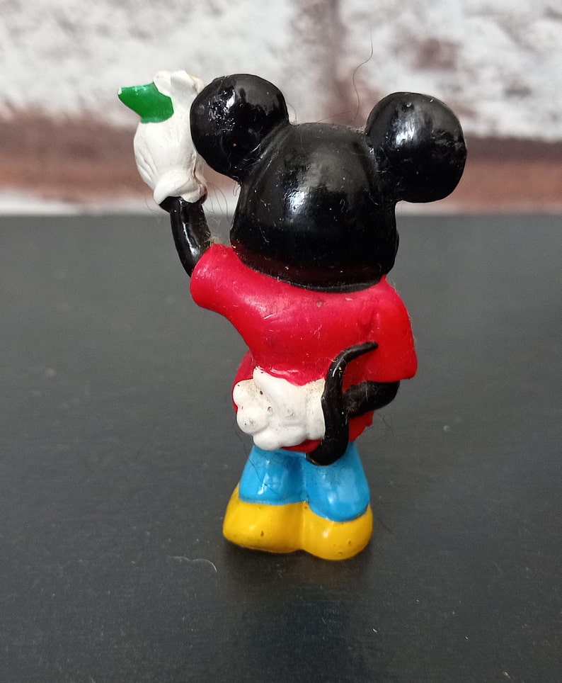 Puede incluir: Una peque&ntilde;a figura de pl&aacute;stico de Mickey Mouse con una camisa roja, pantalones azules y zapatos amarillos. Est&aacute; sosteniendo un guante blanco con una hoja verde en su mano.