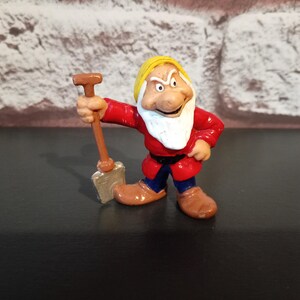 Op de afbeelding: Een beeldje van Grumpy uit Sneeuwwitje, gekleed in een rode tuniek, blauwe broek en bruine schoenen. Hij heeft een witte baard en gele hoed, met een kleine schop. Het beeldje is ongeveer 7,6 cm hoog.
