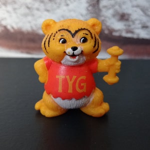 Op de afbeelding: Een kleine, oranje tijgerfiguur met zwarte strepen en een rood shirt. Op het shirt staan de letters "TYG". De tijger houdt een gele halter vast. De figuur staat op een zwart oppervlak.