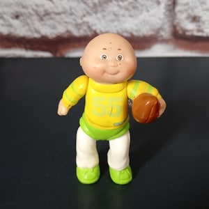 May include: A bald Cabbage Patch Kid doll wearing a yellow and green football uniform with the number 55 on the front. The doll is holding a brown football.
