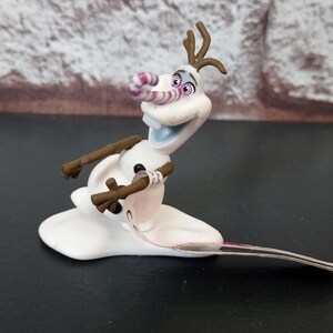 May include: A white Olaf figurine from the movie Frozen, with brown twig arms and hair. The figurine has a pink and white striped nose and is posed on a black surface.