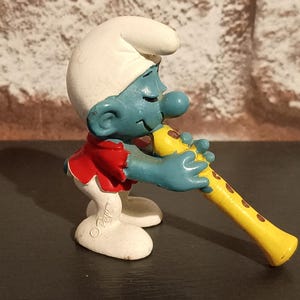 Könnte beinhalten: Eine Schlümpfe-Figur, die eine gelbe Flöte mit braunen Punkten spielt. Der Schlumpf ist blau mit weißer Mütze, rotem Hemd und weißen Schuhen. Die Figur steht auf einer dunklen Oberfläche mit einem ziegelartigen Hintergrund.