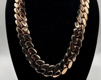 May include: A rose gold chain necklace with a thick, flat curb link design. The necklace is displayed on a black bust, showcasing its polished, reflective surface. The links are oval-shaped and closely connected.