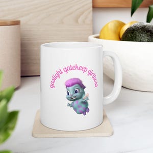 Peut inclure: Mug en céramique blanche avec un personnage de dessin animé bleu et violet et l'inscription "gaslight gatekeep girlboss" en rose. Le mug est posé sur un dessous de verre beige. Idéal pour le café ou le thé.