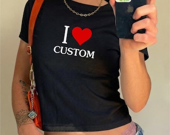 Op de afbeelding: Een zwart T-shirt met korte mouwen en de witte tekst I en CUSTOM met een rood hartsymbool ertussen. Het shirt wordt gedragen met een gouden ketting en een oranje schoudertasriem die aan de linkerkant van het kledingstuk zichtbaar is.