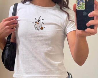 May include: A white short-sleeved t-shirt featuring a central embroidered graphic of a ghost reading an orange book while holding a small pumpkin, surrounded by several small black bats. The shirt is paired with a black shoulder bag.