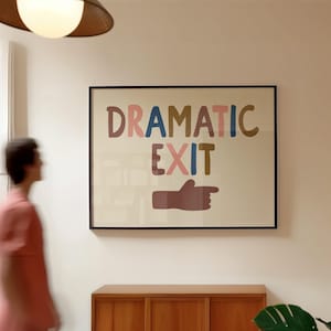May include: A framed art print with the words "DRAMATIC EXIT" in colorful letters. The print includes a brown hand pointing to the right. The artwork is displayed on a white wall above a wooden cabinet.