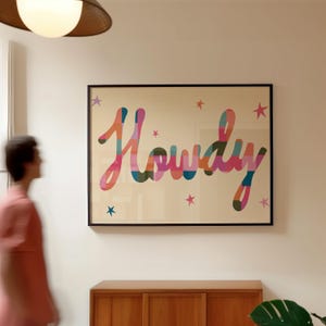 May include: A framed art print featuring the word "Howdy" in vibrant, cursive lettering. The word is made up of stripes of pink, orange, blue, and green. Small pink stars are scattered around the word on a cream background. The print is in a black frame.