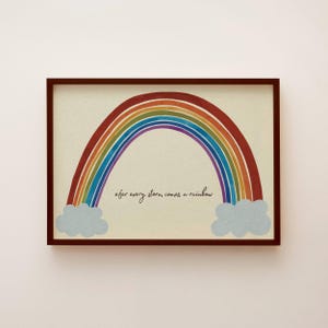 Könnte beinhalten: Gerahmter Druck mit einem bunten Regenbogen und dem Text "after every storm comes a rainbow". Der Regenbogen spannt sich über zwei flauschigen blauen Wolken. Der Rahmen ist dunkelbraun.