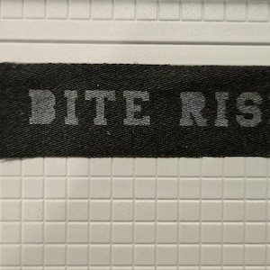 May include: A rectangular black fabric patch with the text BITE RISK printed in a light gray, block-style font. The patch is positioned next to a white ruler, showing a length of approximately 5 inches and a width of approximately 1 inch.