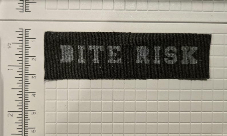 May include: A rectangular black fabric patch with the text BITE RISK printed in a light gray, block-style font. The patch is positioned next to a white ruler, showing a length of approximately 5 inches and a width of approximately 1 inch.