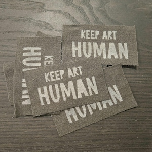 May include: A collection of rectangular, dark grey fabric patches featuring the white printed text KEEP ART HUMAN. The patches are layered on top of each other, displaying the same phrase in a bold, sans-serif font across each individual piece.